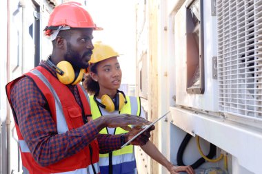 İki endüstriyel Afrikalı mühendis erkek ve kadın güvenlik yeleği ve miğfer giyer, imalat fabrikasında veya lojistik kargo deposunda kontrol düğmesi takarlar. İş yerinde çalışanlar