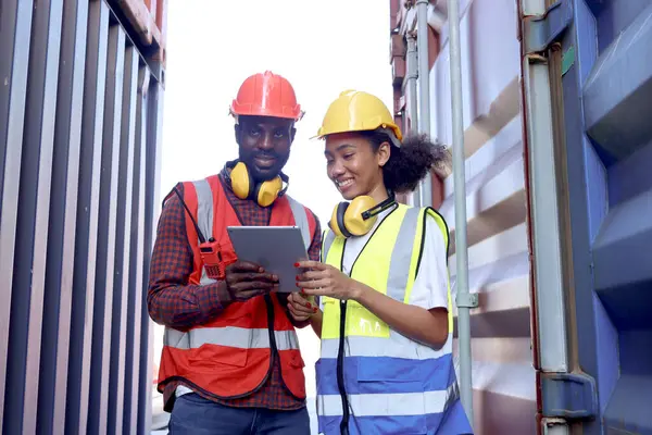 Two African workers wear safety vest and helmet at logistic shipping cargo containers yard. Happy young engineer woman uses digital tablet to discuss results with her male colleague at workplace.