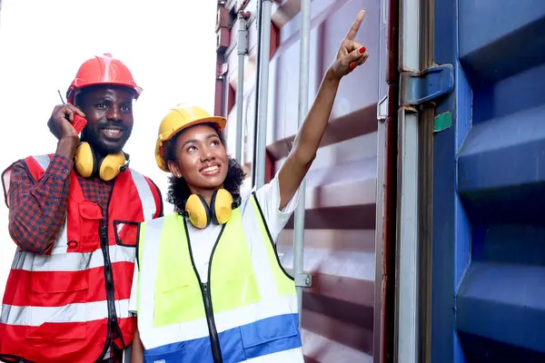 Two African engineer man and woman wearing safety vest and helmet working together at logistic shipping cargo container yard, female worker pointing away at container while male using walkie talkie.