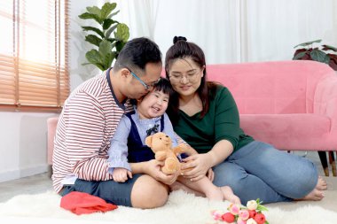 Mutlu tombul Asyalı aile. Baba ve anne oturma odasında küçük kız çocuğuyla oynuyorlar. Ebeveynler çocukla vakit geçirir, sevgi göstermek için sarılırlar. Anne ve babasıyla evde dinlenen bir çocuk..