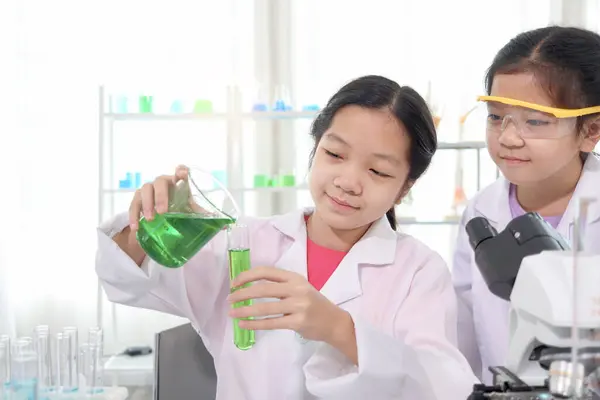 可爱的年轻科学家女学生穿着实验室外套在实验室做科学实验 女学生将烧杯中的绿色试剂溶液倒入试管 儿童学习科学教育 免版税图库照片