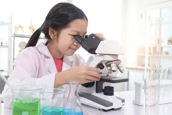 穿着实验室外套的年轻漂亮的女科学家通过显微镜在实验室里研究微生物学 女学生使用实验设备进行科学实验 儿童学习科学教育 图库照片
