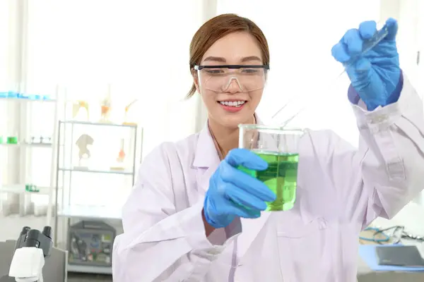 身穿实验室外套 戴着安全眼镜的快乐美丽的亚洲科学家女性的画像做科学实验 研究人员将化学品从管中倒入烧杯用于研究 女性从事科学领域的工作 免版税图库照片