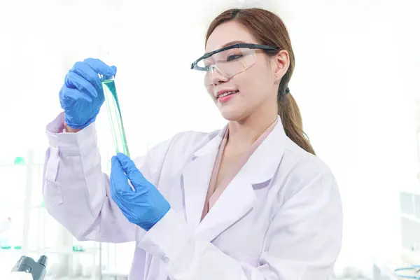 漂亮的亚洲科学家女人 身穿实验室外套 戴着安全眼镜 拿着装有绿色化学物质的试管 研究人员使用仪器进行科学实验研究 妇女在科学领域的工作 图库照片