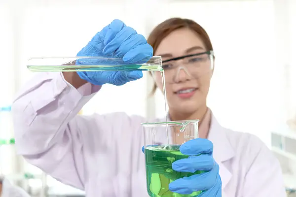 穿着实验室外套的女科学家的手 带着安全眼镜 把化学品从试管倒进烧杯里做科学实验 研究人员使用仪器研究从事科学研究的女性 免版税图库照片