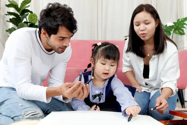 快乐的亚洲家庭 胖胖的小女孩和爸爸妈妈一起在客厅里画画 父母把时间花在与幼儿在家里 妈妈和爸爸一起在家里玩 图库图片