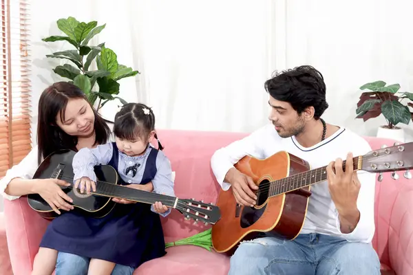 快乐的亚洲家庭 胖胖的小女孩学着和她爸爸一起弹吉他 和父母在一起的孩子在客厅里呆着 孩子和父母在家里玩得很开心 图库图片