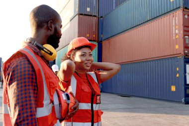 İki mutlu gülümseyen Afrikalı mühendis ve kadın güvenlik yeleği ve miğfer giyer. Lojistik nakliye konteynır deposunda çalışan işçi takımı alacakaranlık günbatımında birlikte çalışırlar..