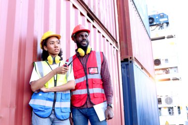Güvenlik yeleği ve miğfer takmış iki mutlu Afrikalı mühendis ve kadının portresi, lojistik kargo konteynır deposunda telsiz ve tabletle dikiliyorlar..