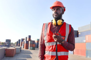 Güvenlik kırmızı yeleği ve miğferi olan, lojistik konteynır nakliye bölümünde çalışan Afrikalı genç mühendis portresi. Elinde telsiz olan ve iş yerinde çalışan erkek mühendis..