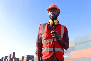 Güvenlik kırmızı yeleği ve miğferi olan, lojistik konteynır nakliye bölümünde çalışan Afrikalı genç mühendis portresi. Elinde telsiz olan ve iş yerinde çalışan erkek mühendis..