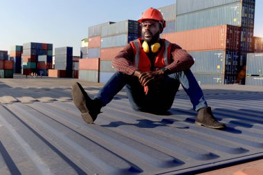 Kırmızı yelek ve kask giyen yorgun Afrikalı genç adam mola sırasında lojistik konteynır nakliye deposunda yerde oturuyor. Erkek mühendis şu anda işyerinde dinleniyor..