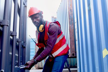 Afro-Amerikalı genç mühendis portresi. Parlak neon renkli yelek ve kask giymiş. Lojistik kargo konteynerinin kapağını açmaya çalışıyor..
