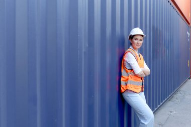Mutlu gülümseyen genç kadın mühendis portresi sarı saçlı, güvenlik yeleği ve beyaz kasklı, güven dolu koluyla lojistik kargo konteynır sahasında duruyor..