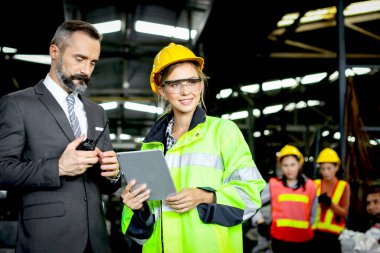 Güzel kadın endüstriyel mühendis kaskı ve koruyucu giysiler giyiyor, tablet tutuyor ve ağır sanayi imalat fabrikasında kıdemli işadamı ile tartışıyor.