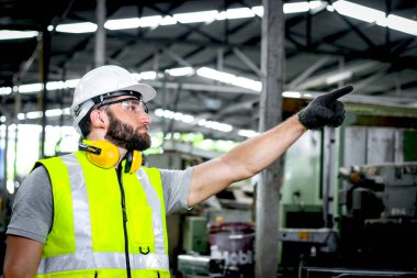 Kask ve güvenlik yeleği giyen erkek sanayi mühendisi fabrika fabrikasını işaret ediyor. İşçi endüstri işyerinde makine ile çalışan.