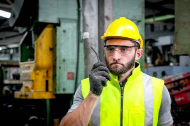 Miğfer ve güvenlik yeleği olan erkek endüstriyel mühendisin portresi. Elinde telsiz var ve fabrikada konuşuyor. Güvenlik gözlüklü işçi endüstri işyerinde çalışıyor..