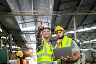 Endüstriyel ustabaşı ve miğfer ve güvenlik yeleği olan işçi imalat fabrikasında tartışırken dizüstü bilgisayar kullanıyorlar. Mühendis işyerini teftiş ediyor ve süreci kontrol ediyor.