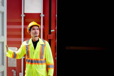 Güvenlik kaskı ve koruyucu giysiler giyen Asyalı mühendis, lojistik nakliye konteynerinde çalışıyor. Erkek işçi ürünleri incelemek için bir konteyner kapısı açıyor..