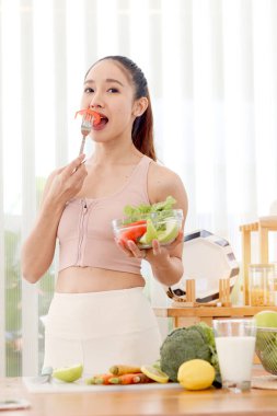 Spor kıyafetli sporcu kız çatal, dilimlenmiş domates ve salata kasesi tutuyor. Mutfaktaki güzel mutlu Asyalı kadın sağlıklı sebze yiyor. Zayıflıyor ve zayıflıyor. Sağlıklı gıda sporu.
