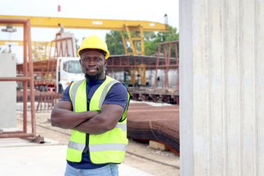 Güvenlik yeleği ve miğferi olan mutlu gülümseyen Afrikalı mühendis portresi inşaat alanında kolları bağlı duruyor. İnşaat alanının zemin katında çalışan genç mimar.