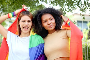 Mutlu gülümseyen eşcinsel lezbiyen çift ellerinde gökkuşağı bayrağı tutuyor. Güzel bir kadın ve Afrikalı kıvırcık saçlı kız arkadaşı, LGBT aşığı ve eşcinsel gurur hareketi kavramını birlikte kutluyorlar.