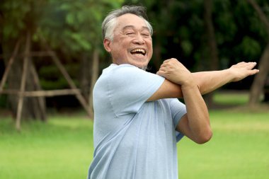 Yaşlı Asyalı adam egzersiz yapmadan önce vücudunu esnetiyor ve sabah parkta koşuyor. Yeşil bahçede iyi son sınıf egzersizleri. Doğanın arka bahçesinde aktif sağlıklı yaşlılar egzersizi..