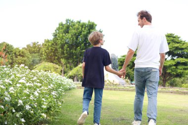 Baba ve oğul parkta yürürken el ele tutuşuyorlar. Mutlu aile, ebeveyn ve çocuk tatilde güzel bir bahçede birlikte vakit geçiriyorlar. Küçük çocuk dışarıda babasıyla oynuyor..