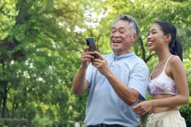 Mutlu Asyalı adam ve güzel kız yeşil yaz bahçesinde selfie çekiyorlar. Dedem parkta genç torunuyla dışarıda vakit geçirmekten hoşlanır. Üst düzey erkek cep telefonu kullanıyor..