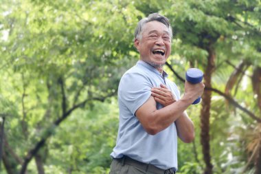 Yeşil halk parkında yaşlı mutlu Asyalı adam dambılı kaldırıyor. Neşeli son sınıf öğrencisi bahçede kas gücü için egzersiz yapmaktan hoşlanıyor. Çevresi doğa ile çevrili aktif sağlıklı yaşlı insanlar..