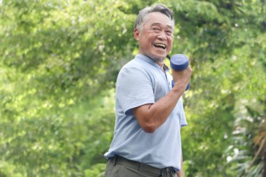 Yeşil halk parkında yaşlı mutlu Asyalı adam dambılı kaldırıyor. Neşeli son sınıf öğrencisi bahçede kas gücü için egzersiz yapmaktan hoşlanıyor. Çevresi doğa ile çevrili aktif sağlıklı yaşlı insanlar..