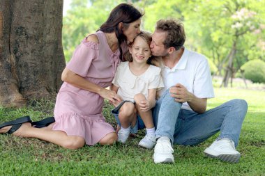 Bahçenin dışında güzel, mutlu bir aile. Yeşil Park 'ta ağaçların altında otururken neşeli ebeveynler kızlarını öpüyor. Baba, anne ve çocuk birlikte dışarıda vakit geçiriyorlar..