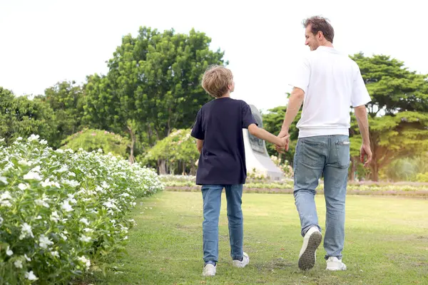 Baba ve oğul parkta yürürken el ele tutuşuyorlar. Mutlu aile, ebeveyn ve çocuk tatilde güzel bir bahçede birlikte vakit geçiriyorlar. Küçük çocuk dışarıda babasıyla oynuyor..