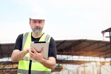 Güvenlik yeleği ve miğferi olan kıdemli mühendis inşaat alanında bilgi kontrol etmek için tablet tutuyor. Yaşlı mimar inşaat sahasının zemin katında çalışıyor. Olgun erkek işçi.