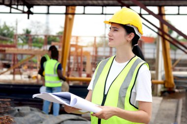 Güvenlik yeleği ve miğferi olan genç ve ciddi mühendis kadın mühendisin yan görünüşü inşaat alanının planlarını içeriyor. İnşaat alanında çalışmayı düşünen kadın mimara konsantre ol.