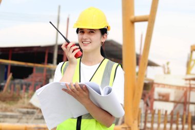 Güvenlik yeleği ve miğferi olan genç ve mutlu mühendis kadın mühendis inşaat alanında plan ve telsiz tutuyor. İnşaat alanında çalışan gülümseyen kadın mimar..