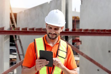 Güvenlik yeleği ve miğferi olan mutlu işçi mühendisinin portresi inşaat alanında incelenirken bilgiyi kontrol etmek için dijital tablet içeriyor. İnşaat alanında çalışan genç mimar.