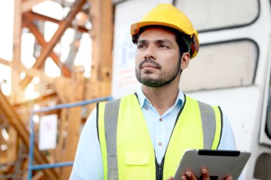 Güvenlik yeleği ve miğferi olan portre yakışıklı mühendis inşaat alanını incelerken dijital tablet tutuyor. Genç mimar inşaat alanında çalışıyor..