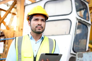 Güvenlik yeleği ve miğferi olan portre yakışıklı mühendis inşaat alanını incelerken dijital tablet tutuyor. Genç mimar inşaat alanında çalışıyor..