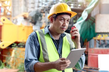 Güvenlik yeleği ve miğferi olan kıdemli Asyalı işçi mühendisi inşaat alanında inceleme yaparken bilgiyi kontrol etmek için dijital tablet tutuyor. İş yerinde çalışan yaşlı, olgun bir mimar..