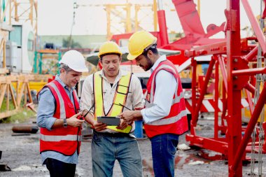 Kıdemli mühendis ve güvenlik yeleği ve miğfer saklama tabletli işçi grupları inşaat alanında tartışıyorlar. Olgun yaşlı ve genç mimar inşaat alanında birlikte çalışıyor.