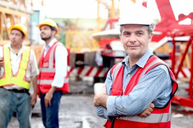 Güvenlik yeleği ve miğferi olan ve inşaat alanında çalışan iş arkadaşıyla yan yana duran mutlu kıdemli mühendis ustabaşının portresi. İnşaat alanında olgun yaşlı bir mimar çalışıyor.