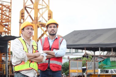 Güvenlik yeleği ve miğferi olan iki işçinin portresi kollarını kavuşturup şantiyede tartışıyorlar. Yaşlı mühendis ve genç işçiler şantiyede birlikte çalışıyorlar.. 