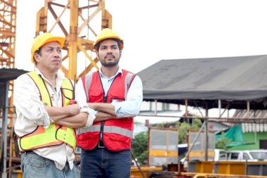 Güvenlik yeleği ve miğferi olan iki işçinin portresi kollarını kavuşturup şantiyede tartışıyorlar. Yaşlı mühendis ve genç işçiler şantiyede birlikte çalışıyorlar.. 