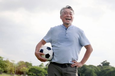 Yeşil halk parkında futbol oynayan mutlu Asyalı adam. Neşeli son sınıf öğrencisi en sevdiği futbol sporuyla dışarıda egzersiz yapmayı seviyor. Bahçenin arka bahçesinde aktif sağlıklı yaşlılar egzersizi.