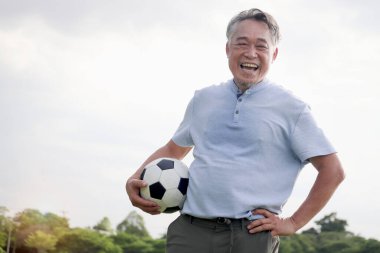 Yeşil halk parkında futbol oynayan mutlu Asyalı adam. Neşeli son sınıf öğrencisi en sevdiği futbol sporuyla dışarıda egzersiz yapmayı seviyor. Bahçenin arka bahçesinde aktif sağlıklı yaşlılar egzersizi.