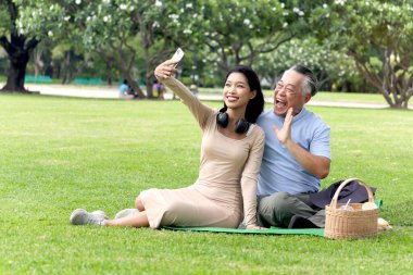Mutlu aile, yaşlı Asyalı bir adam ve güzel bir kız yeşil yaz bahçesinde piknik yaparken selfie çekmek için cep telefonu kullanıyor. Büyükbaba dışarıda torunuyla parkta vakit geçiriyor.. 