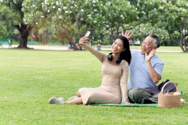 Mutlu aile, yaşlı Asyalı bir adam ve güzel bir kız yeşil yaz bahçesinde piknik yaparken selfie çekmek için cep telefonu kullanıyor. Büyükbaba dışarıda torunuyla parkta vakit geçiriyor.. 