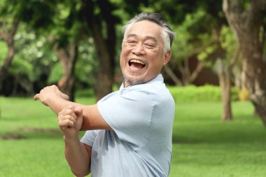 Yaşlı Asyalı adam egzersiz yapmadan önce vücudunu esnetiyor ve sabah parkta koşuyor. Yeşil bahçede iyi son sınıf egzersizleri. Doğanın arka bahçesinde aktif sağlıklı yaşlılar egzersizi..