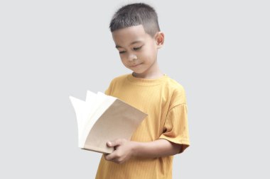 Sarı tişörtlü küçük bir Asyalı çocuk gri arka planda izole bir kitap tutuyor. Genç öğrenci okumayı sever. Okul kitabıyla portre çocuğu. Çocuk konsepti eğitimi.